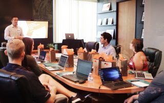 A group of experts sitting at a conference room table with laptops and listening to a presenter